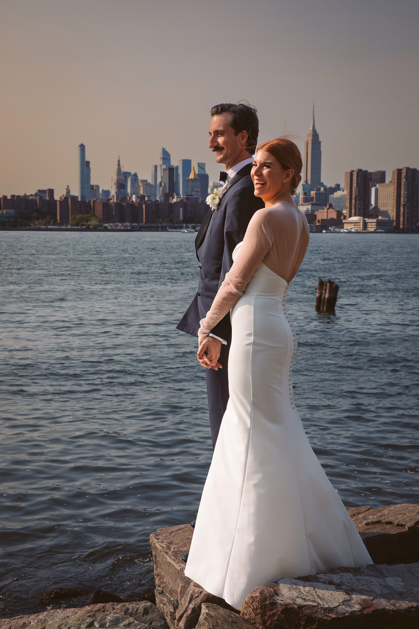 A bride and groom laughing in front of the New York City skyline during their Wythe Hotel wedding