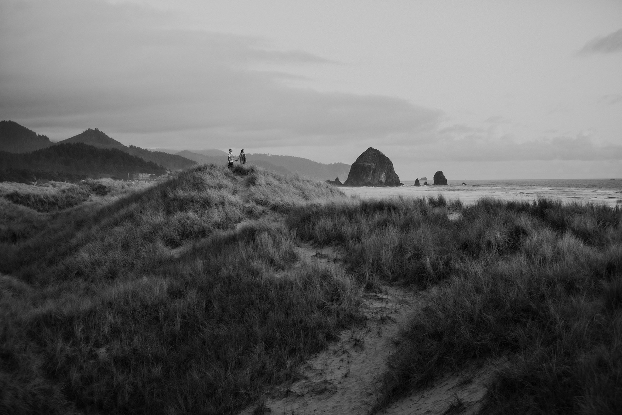 A black and white photo of a couple standing in the sane dunes on Cannon Beach after their surprise proposal