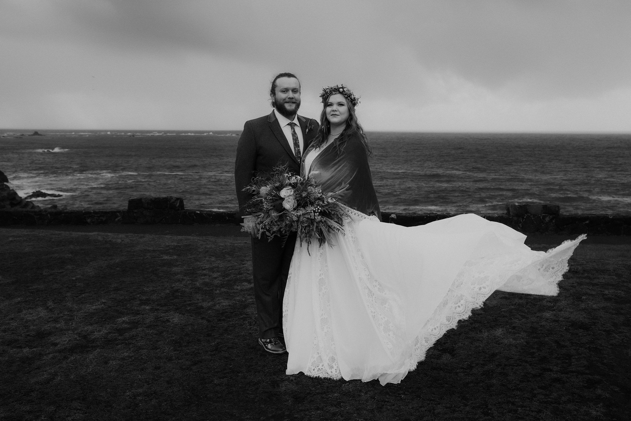 A black and white photo of a couple on a cliffside in Oregon with the brides dress blowing in the wind