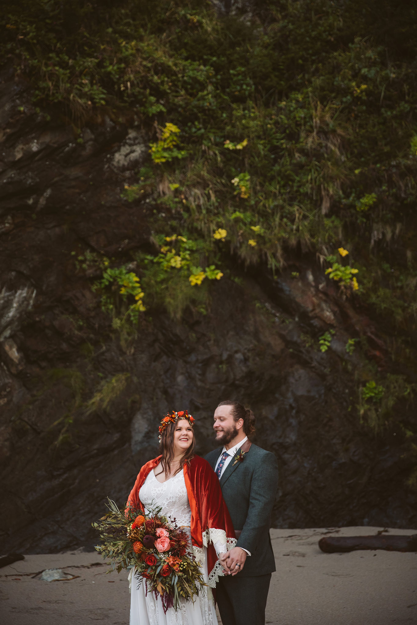 A wedding portrait of a bride and groom on the beach with a moss covered rock wall in the background